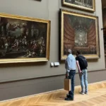 Museum visitors observing grand historical paintings in a gallery of the Louvre Museum